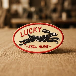 May include: An oval-shaped embroidered patch with a cream-coloured background and a red border. It features a black running rabbit graphic with white accents, the red embroidered text "LUCKY" above, and the red embroidered text "STILL ALIVE" below.