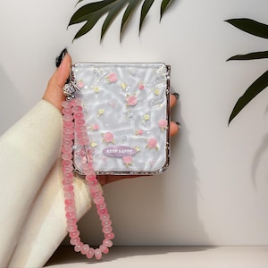 May include: A square, white, textured compact mirror case featuring a pattern of small pink roses and yellow flowers. A purple label with the text "keep happy" is centered near the bottom. A long, pink, translucent beaded wrist strap is attached.