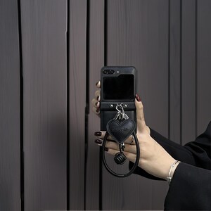 May include: A black smartphone case featuring a horizontal strap with two silver metal rings. Attached to the rings is a black heart-shaped charm embossed with the text "sullan-life" and a matching black circular wrist strap with a knotted detail.