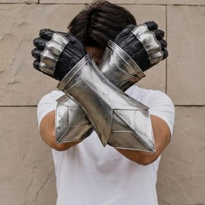 May include: A pair of silver metal gauntlets featuring articulated finger plates and a textured black leather base. The gauntlets extend to cover the forearms with a polished, angular steel design, worn crossed over the chest in a protective stance.