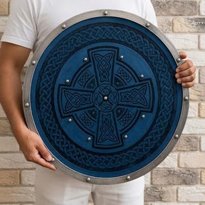 May include: A circular shield featuring a deep blue surface with a black embossed Celtic cross and intricate knotwork patterns. The shield has a metallic silver rim secured with evenly spaced rivets around the outer edge and centre.