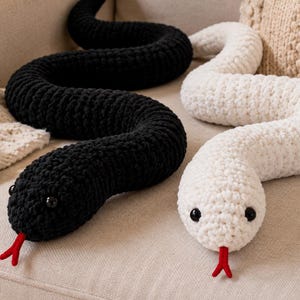 Peut inclure: Deux longs jouets serpents en crochet, l'un noir uni et l'autre blanc uni. Chaque serpent présente une tête arrondie avec deux petits yeux en perles noires et une langue bifide en fil rouge sortant de la bouche. Les corps sont texturés.