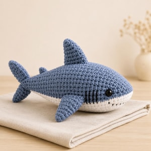 May include: A blue and white crocheted shark plush toy featuring a prominent dorsal fin, a tail fin, side fins, and black embroidered gill slits. The shark has a rounded body with a white underbelly and a single black plastic eye visible.