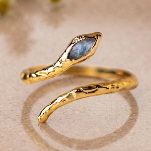 May include: A gold-toned open ring shaped like a coiled snake with a textured, hammered metal surface. The head of the snake features a single, elongated, marquise-cut blue stone set within the metal, creating a distinct focal point.
