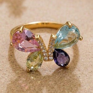 May include: A gold ring featuring a butterfly design composed of four pear-shaped gemstones in pink, light blue, light green, and dark purple. The center of the butterfly is accented with small clear stones set in a vertical gold bar.