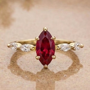 Pode incluir: Um anel de ouro com uma pedra preciosa vermelha central em lapidação marquise, presa por quatro garras. A banda é adornada em cada lado por duas pedras transparentes menores em lapidação marquise, criando um design simétrico com um acabamento metálico polido.