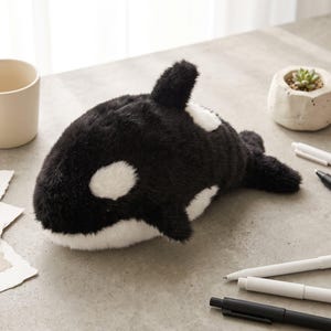 May include: A plush orca toy featuring a soft black and white faux fur exterior, a dorsal fin, and characteristic white eye patches. The stuffed animal is positioned horizontally, showcasing its rounded body shape and detailed plush texture.