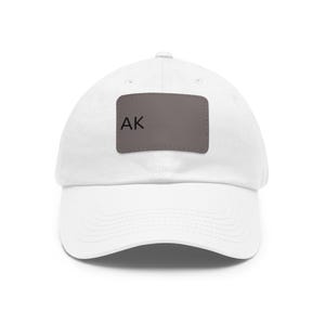 Op de afbeelding: Een witte baseballpet met een rechthoekige grijze patch in het midden van het voorpaneel. De patch bevat de zwarte tekst AK aan de linkerkant. De pet heeft een gebogen klep, een knoop aan de bovenkant en gestikte oogjes voor ventilatie.