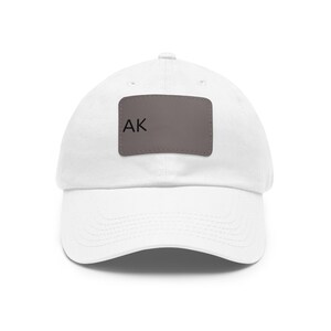Op de afbeelding: Een witte baseballpet met een rechthoekige grijze patch in het midden van het voorpaneel. De patch bevat de zwarte tekst AK aan de linkerkant. De pet heeft een gebogen klep, een knoop aan de bovenkant en gestikte oogjes voor ventilatie.