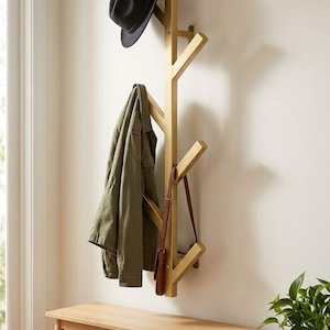 Pode incluir: Um cabide de parede dourado em forma de ramo de árvore com vários ganchos. Um chapéu fedora preto está pendurado no gancho superior, enquanto um casaco verde-azeitona e uma pequena bolsa de ombro de couro castanho pendem dos ramos inferiores.