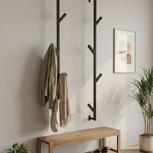Puede incluir: Dos percheros verticales de pared hechos de metal negro con ganchos en forma de rama. Uno sostiene una chaqueta verde claro, una bufanda beige y una bolsa de tela. Debajo hay un banco de madera con varios pares de zapatos.