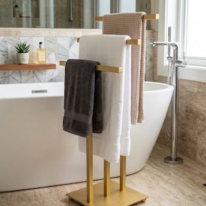 Puede incluir: Un toallero de metal dorado con tres barras horizontales a diferentes alturas, apoyado sobre una base cuadrada. El soporte sostiene una toalla gris oscuro, una toalla blanca y una toalla beige con textura de panal de abeja.