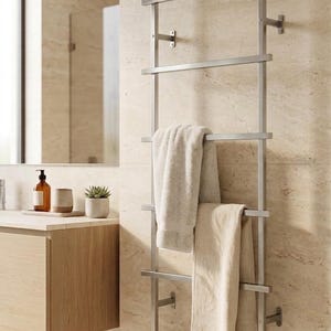 May include: A wall-mounted, brushed metal towel rack featuring a ladder-style design with six horizontal bars. Two beige cotton towels are draped over the middle and lower rungs of the vertical frame, which is secured to the wall with metal brackets.