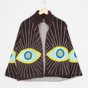 Puede incluir: Una chaqueta abierta de color marrón oscuro con grandes motivos de ojos bordados en el frente y las mangas. Los ojos tienen iris azul claro, pupilas grises y contornos amarillos con líneas blancas radiantes. El forro interior es blanco con finas rayas negras.
