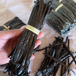 Könnte beinhalten: Ein Bündel dunkelbrauner, länglicher Vanilleschoten, die mit einer hellen Bastkordel zusammengebunden sind. Weitere Bündel von Vanilleschoten, teilweise in Klarsichtfolie verpackt, sind um das zentrale, gebundene Bündel herum verteilt.