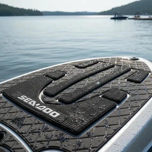 Puede incluir: Una alfombrilla de tracción negra Sea-Doo con textura de patrón de diamante y secciones de agarre rectangulares elevadas. La alfombrilla presenta el logotipo blanco de Sea-Doo impreso en la superficie, cubierta de gotas de agua.