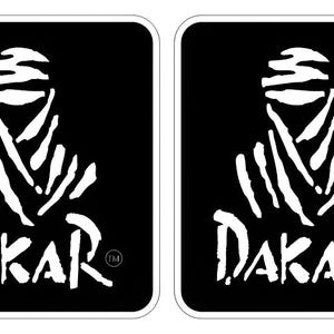 May include: Two rectangular black stickers with rounded corners, each featuring a white graphic of a stylized person wearing a head wrap above the word DAKAR in a bold, brush-stroke font, with a small trademark symbol next to the text.