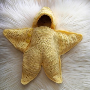 May include: A bright yellow crochet star-shaped bunting with a hood. The garment features a five-pointed design with a central opening for the head and two small yellow buttons on one of the upper points to secure the fabric around the body.