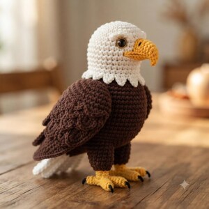 May include: A crocheted bald eagle figurine featuring a white head, a dark brown body, and a yellow beak and feet. The eagle has a textured, stitched appearance with a rounded head, dark eyes, and layered wing details crafted from yarn.