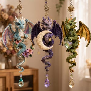 May include: Three hanging crochet dragon ornaments suspended by metal chains. The first is light blue with pink flowers, the second is purple with a cream crescent moon, and the third is green holding a clear crystal sphere, each with a crystal drop.