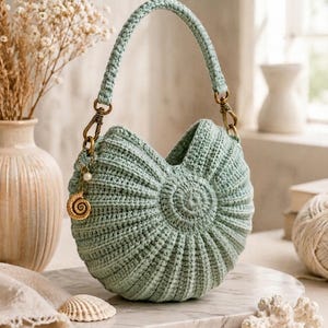 May include: A sage green crochet handbag with a rounded, shell-like ribbed texture and a single handle. The bag features a gold-toned metal clasp on one side, adorned with a small white pearl bead and a gold spiral seashell charm hanging from it.
