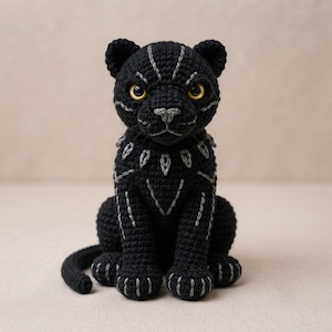 May include: A black crocheted panther figurine featuring bright yellow eyes and silver-grey embroidered detailing on the face, chest, and paws. The stuffed animal is in a seated position with its tail curled forward, showcasing intricate yarn work.