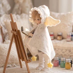 May include: A ball-jointed doll with blonde hair wearing a white hooded cape with ruffled trim, angel wings, and star-shaped accents. The doll stands before a wooden easel, holding a small spiral-bound sketchbook. The cape features a subtle moon and star pattern.