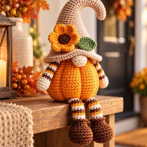 Può includere: Una statuetta di gnomo all'uncinetto con un cappello a punta color marrone chiaro decorato con un girasole giallo e una foglia verde. Il corpo è una zucca arancione rotonda con braccia e gambe a righe bianche, arancioni e marroni.