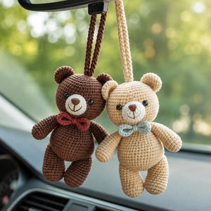 May include: Two crocheted teddy bear ornaments hanging from a rearview mirror. One bear is dark brown with a red bow tie, and the other is light beige with a light blue bow tie. Both bears feature stitched facial details and braided hanging loops.