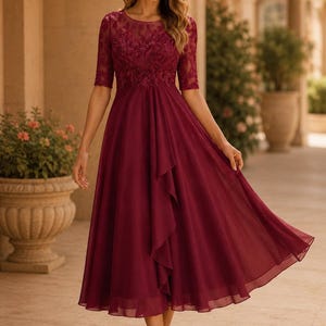 May include: A burgundy midi dress featuring a round neckline, elbow-length lace sleeves, and a sheer lace bodice with intricate embroidery. The skirt is made of flowing chiffon fabric with a vertical ruffled detail cascading down the front side.