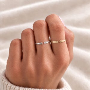 May include: Two open-style rings worn on adjacent fingers. One ring is silver-toned and features the letters O and K engraved on its ends. The second ring is gold-toned and features the letters N and R engraved on its ends. Both rings are thin.