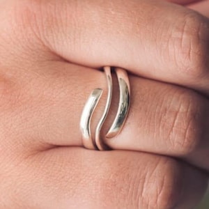 Puede incluir: Un anillo plateado pulido con un diseño de bypass que presenta dos bandas lisas y curvas que se envuelven alrededor del dedo. El metal tiene un acabado reflectante de alto brillo con bordes redondeados, creando una forma fluida y continua.
