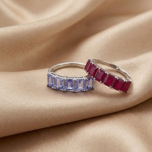 May include: Two silver-toned rings featuring a row of rectangular-cut gemstones. One ring is set with light blue stones, while the other ring is set with deep red stones. Both rings have a polished metal band and a prong setting for each stone.