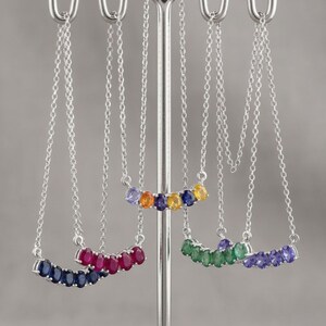 Può includere: Quattro collane in argento appese a un espositore in metallo. Ogni collana presenta una barra curva incastonata con cinque pietre preziose dal taglio ovale in diversi colori, tra cui blu, rosso, verde e una combinazione multicolore di giallo, arancione e viola.