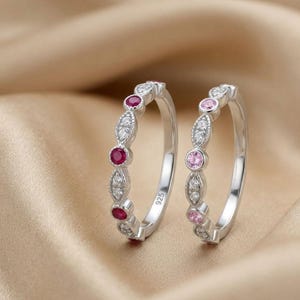 Könnte beinhalten: Zwei Silberringe mit 925er-Stempel, die jeweils eine Reihe abwechselnder runder und marquiseförmiger Fassungen aufweisen. Ein Ring ist mit dunkelroten und klaren Steinen besetzt, der andere mit hellrosa und klaren Steinen.