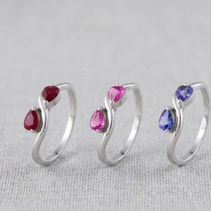 Könnte beinhalten: Drei silberfarbene Ringe, jeder mit einer Bypass-Schiene und zwei birnenförmigen Edelsteinen. Der erste Ring hat tiefrote Steine, der zweite leuchtend rosa Steine und der dritte kräftig blaue Steine, alle gefasst in polierten Metallschienen.