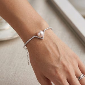 Puede incluir: Una pulsera de tono plateado con un centro en forma de V adornado con pequeñas piedras transparentes y una perla redonda de color rosa pálido. La banda está incrustada con diminutas piedras transparentes e incluye una cadena de extensión.