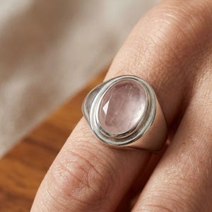 Puede incluir: Un anillo de sello de plata con una gran piedra preciosa de cuarzo rosa de forma ovalada y facetada en el centro. La banda de metal tiene un acabado liso y pulido, y la piedra muestra un tono rosa translúcido con facetas visibles.