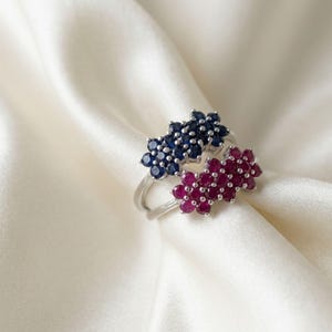 May include: Two silver rings featuring cluster settings. One ring is adorned with a cluster of round dark blue sapphire gemstones, while the other ring features a cluster of round deep pink ruby gemstones, both set in polished silver bands.