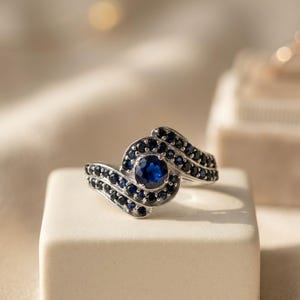May include: A silver ring featuring a central round-cut blue sapphire surrounded by a halo of smaller blue sapphires. The band has a bypass design with two curving rows of pavé-set blue sapphires that extend from the centre stone along the shank.