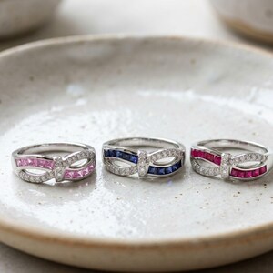 Op de afbeelding: Drie zilverkleurige ringen met een oneindigheidsontwerp. Elke ring is versierd met een rij kleine heldere stenen en een rij vierkant geslepen gekleurde stenen, in roze, donkerblauw en rood, gerangschikt in een symmetrisch patroon.