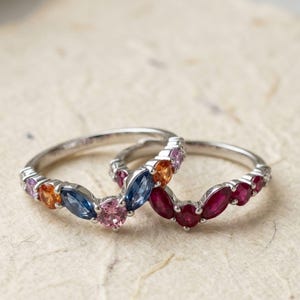 Peut inclure: Deux bagues argentées au design incurvé en forme de V. L'une est ornée de pierres précieuses taille marquise et ronde dans des tons rose, orange et bleu. La seconde est sertie de pierres précieuses rouge foncé taille marquise et ronde.
