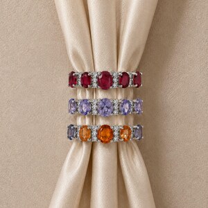 Puede incluir: Tres anillos apilados con piedras preciosas de corte ovalado en rojo, morado claro y naranja, acentuados con pequeñas piedras transparentes en una banda de metal plateado. Los anillos están dispuestos verticalmente con piedras de colores.