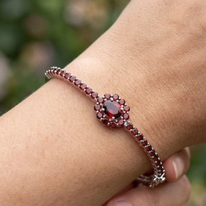 Könnte beinhalten: Ein silberfarbenes Armband mit einer durchgehenden Reihe kleiner, runder, tiefroter Granatsteine. In der Mitte des Armbands befindet sich ein größerer, ovaler Granat, der von einer Gruppe kleinerer, runder Granatsteine in Blütenform umgeben ist.