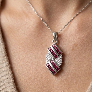 May include: A silver pendant necklace featuring a geometric design with channel-set square rubies and small pavé-set diamonds. The pendant is suspended from a delicate silver chain, showcasing a structured, layered arrangement of red gemstones.