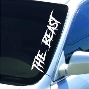 May include: A white vinyl decal applied to the corner of a car windshield. The decal features the text THE BEAST written in a jagged, distressed font style that mimics claw marks or sharp, torn edges, positioned diagonally along the glass edge.
