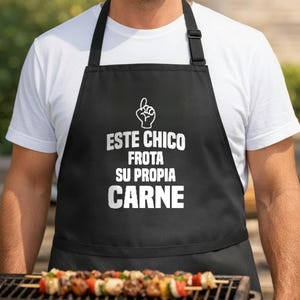 May include: A black apron featuring a white graphic of a hand with an index finger pointing up, above the white text that reads: ESTE CHICO FROTA SU PROPIA CARNE. The apron has black neck and waist straps and is worn over a plain white t-shirt.