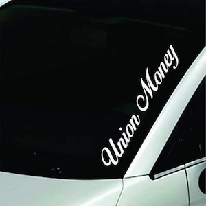 May include: A white vinyl decal applied to the corner of a vehicle windshield. The decal features the text Union Money written in a stylized, cursive script font, positioned diagonally along the edge of the glass surface of the car.