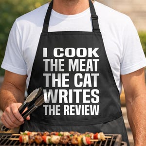 May include: A black apron featuring the white text I COOK THE MEAT THE CAT WRITES THE REVIEW. The apron has black neck and waist straps with an adjustable buckle. The design is centered on the chest area of the garment, providing full coverage.