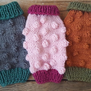 May include: Three knitted dog sweaters in different colours. The sweaters have a textured, bobble stitch pattern and contrasting coloured trim around the neck and bottom. The sweaters are grey with green trim, pink with burgundy trim, and orange with olive green trim.